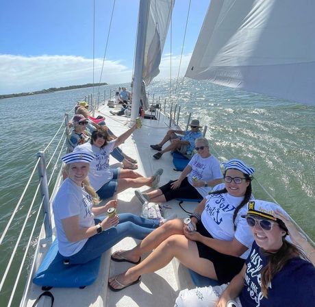 Group of women relaxing on a sailboat in sunny coastal waters, wearing sailor hats and a visible 'bride' cap, holding drinks with white sails billowing and shoreline in the distance.