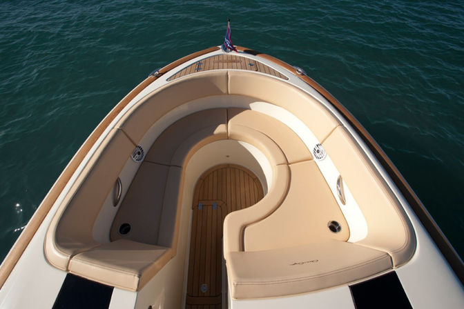 Top-down view of a boat bow with tan curved cushioned seating, teak deck flooring, stainless cup holders and small flag, floating on deep blue-green water