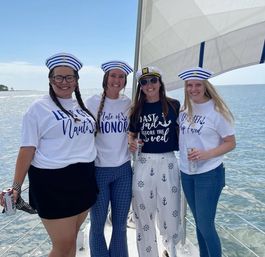 Four women in sailor hats and nautical tees smiling on a sailboat over calm coastal waters, celebrating a bachelorette 'last sail before the veil'.