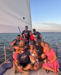 Friends on a sailboat at sunset on a calm coastal bay — smiling group in summer outfits and sunglasses holding drinks beneath a large white sail.