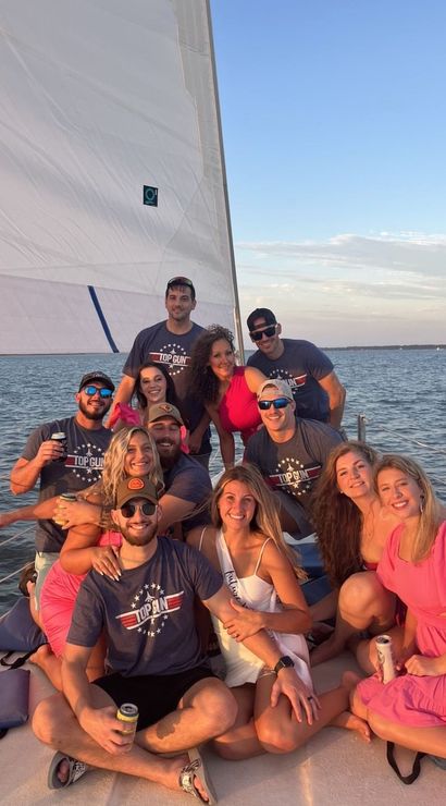 Friends on a sailboat at sunset on a calm coastal bay — smiling group in summer outfits and sunglasses holding drinks beneath a large white sail.