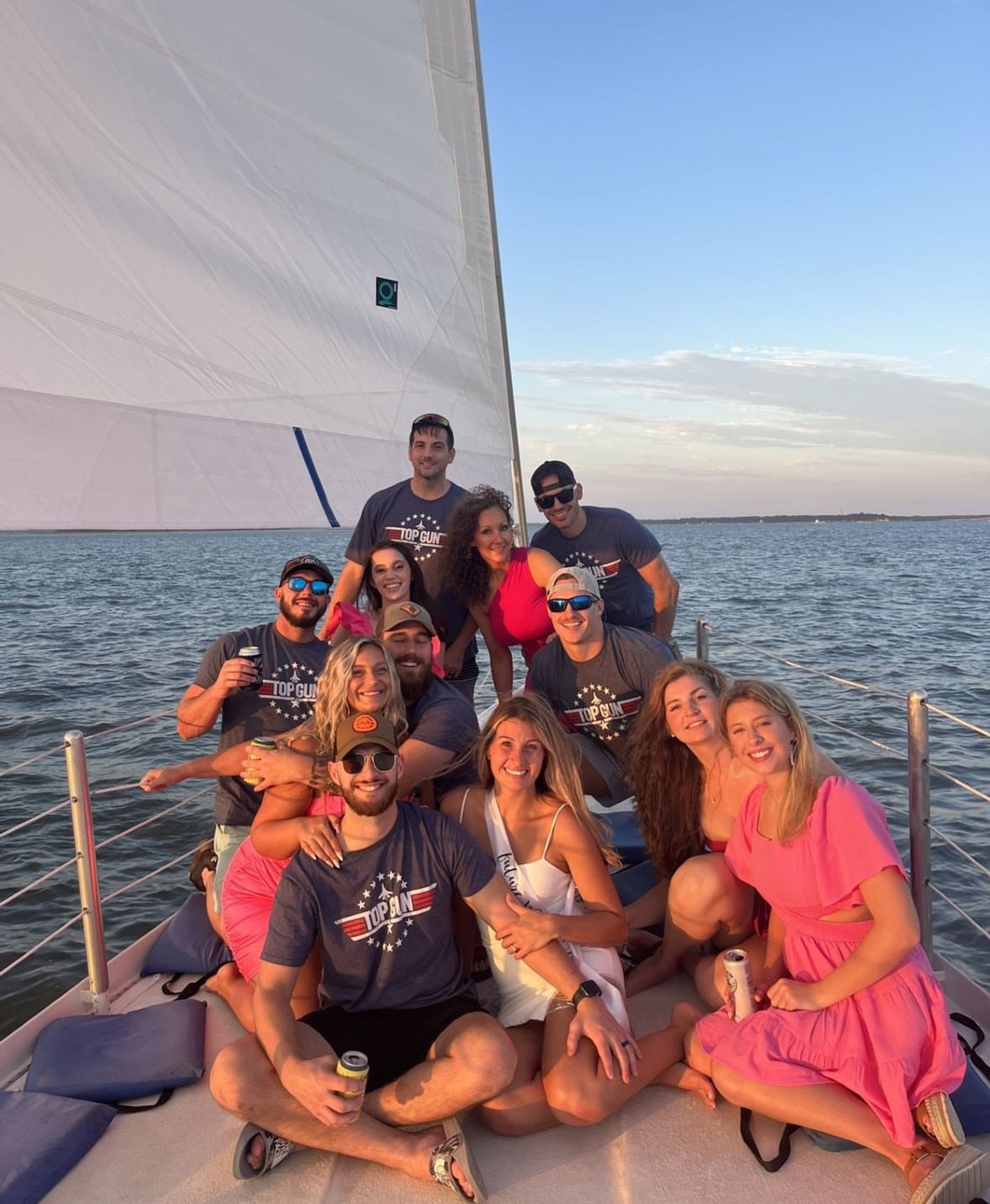 Friends on a sailboat at sunset on a calm coastal bay — smiling group in summer outfits and sunglasses holding drinks beneath a large white sail.