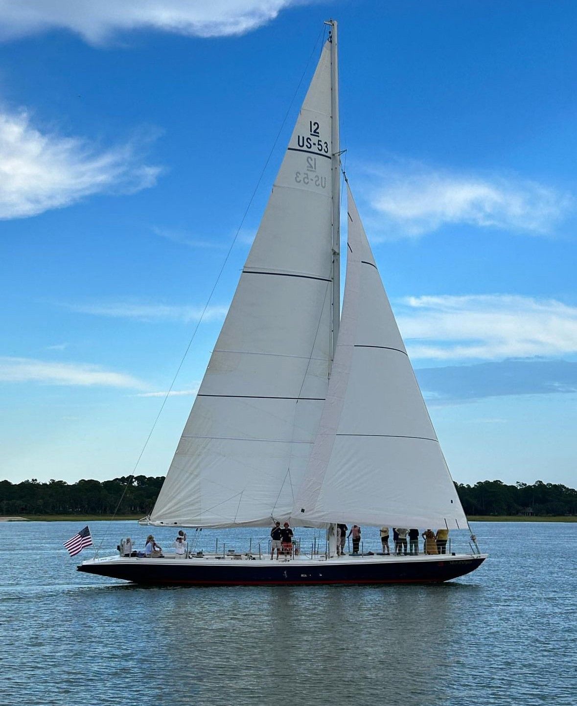 Large classic sailboat with tall white sails and an American flag, passengers on deck, gliding across a calm coastal bay with a tree-lined shore under a bright blue sky.