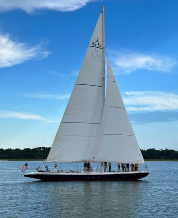 Large classic sailboat with tall white sails and an American flag, passengers on deck, gliding across a calm coastal bay with a tree-lined shore under a bright blue sky.