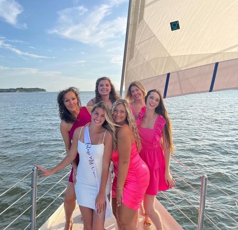 Six smiling women in pink and white dresses posed on a sailboat deck with a coastal bay, blue sky, and warm golden-hour light during a summer cruise.