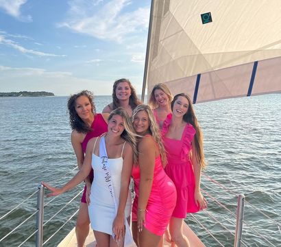 Six smiling women in pink and white dresses posed on a sailboat deck with a coastal bay, blue sky, and warm golden-hour light during a summer cruise.