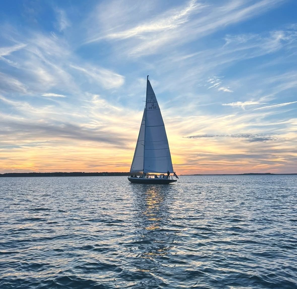 Sailboat gliding on calm coastal waters at sunset, tall white sail silhouetted against a pastel sky with rippled reflections on the sea.