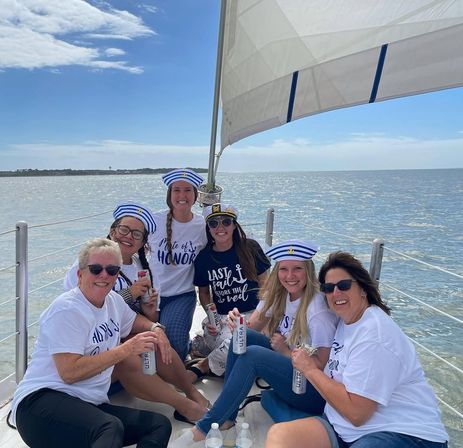 Group of six women wearing striped sailor hats and matching bachelorette-themed shirts, smiling and holding drinks while seated on a sailboat under full sail on a sunny coastal bay.