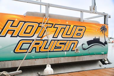 Bright orange and teal "Hot Tub Cruisin" graphic on a pontoon boat at a marina, with palm tree and wave icons, dock rope and fender visible.