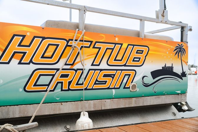 Bright orange and teal "Hot Tub Cruisin" graphic on a pontoon boat at a marina, with palm tree and wave icons, dock rope and fender visible.