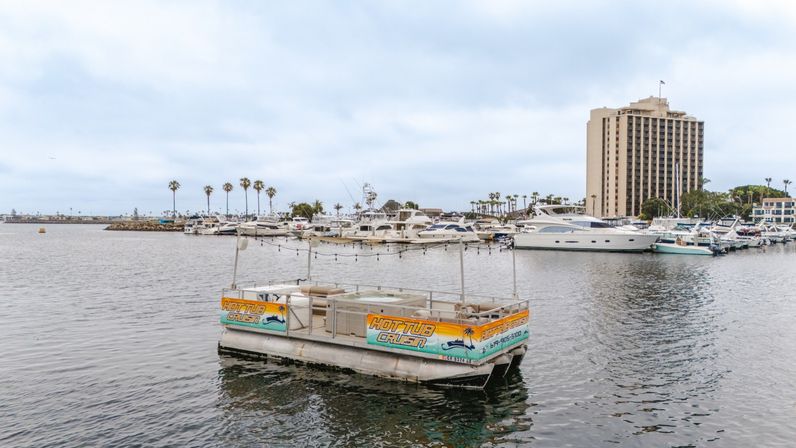 Floating hot-tub platform with colorful signage and string lights in a coastal marina, surrounded by moored yachts, palm trees and a tall waterfront hotel under a cloudy sky