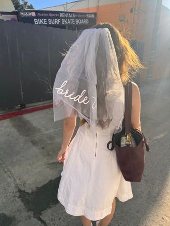 Back view of a bachelorette in a white dress wearing a 'bride' veil and carrying a bag, walking through a sunlit urban alley with a bike/skate rental sign overhead.
