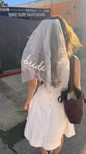 Back view of a bachelorette in a white dress wearing a 'bride' veil and carrying a bag, walking through a sunlit urban alley with a bike/skate rental sign overhead.
