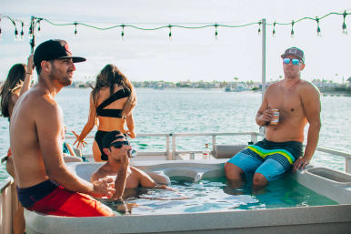 Boat party: people in swimwear relaxing in a hot tub on a pontoon boat, sipping canned drinks with a sunny harbor and string lights in the background.