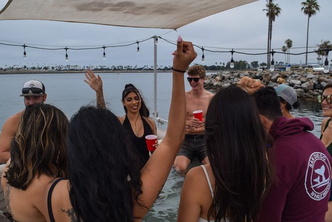Group of friends partying on a covered boat at a coastal California marina, raising hands and holding red cups under string lights with palm trees and rocky shoreline in the background.