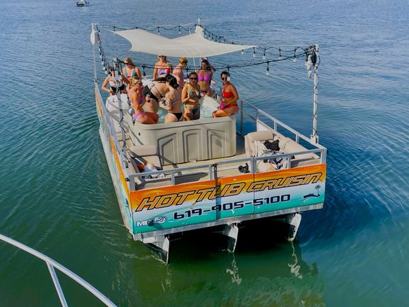 Group of adults in swimsuits relaxing and partying in a shaded hot tub on a colorful pontoon boat floating on calm coastal waters.