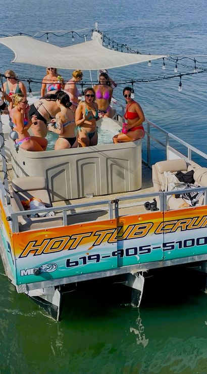 Group of adults in swimsuits relaxing and partying in a shaded hot tub on a colorful pontoon boat floating on calm coastal waters.