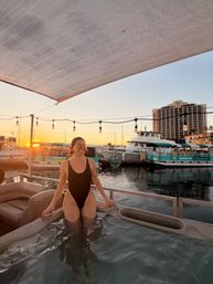 Smiling person in a black one-piece swimsuit relaxing in a boat hot tub at a marina at sunset, string lights overhead and yachts docked along the calm harbor