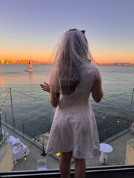 Bride-to-be in a veil on a balcony overlooking a waterfront sunset with a sailboat, twinkling string lights and a distant city skyline