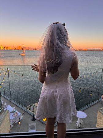Bride-to-be in a veil on a balcony overlooking a waterfront sunset with a sailboat, twinkling string lights and a distant city skyline