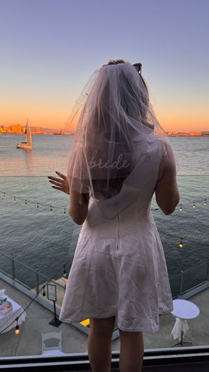 Bride-to-be in a veil on a balcony overlooking a waterfront sunset with a sailboat, twinkling string lights and a distant city skyline