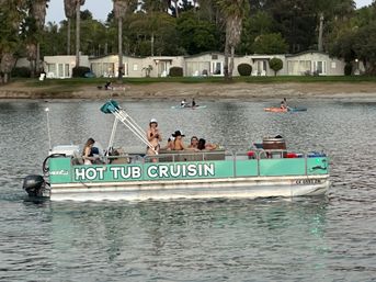 People enjoying a turquoise pontoon hot tub near paddleboarders on a calm bay with palm-lined shoreline and beachfront cottages at dusk.