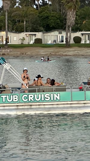 People enjoying a turquoise pontoon hot tub near paddleboarders on a calm bay with palm-lined shoreline and beachfront cottages at dusk.