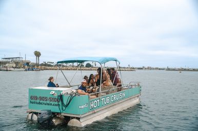 Teal pontoon party boat with canopy carrying a group of friends lounging and chatting on a calm coastal marina lined with palm trees