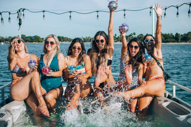 Six friends in colorful swimsuits at a summer boat party, laughing and splashing water while holding glittery drink tumblers on a sunny tree-lined waterfront with string lights overhead.