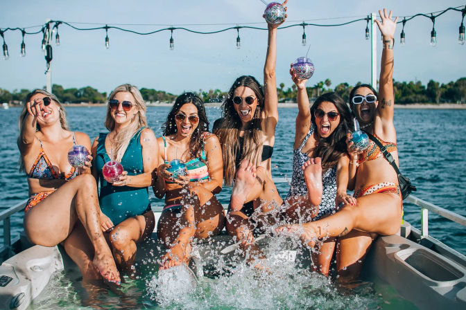 Six friends in colorful swimsuits at a summer boat party, laughing and splashing water while holding glittery drink tumblers on a sunny tree-lined waterfront with string lights overhead.