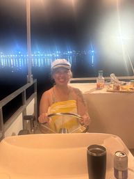 Smiling person in a captain’s hat steering a boat at night with marina lights reflecting on calm harbor water and a snack table nearby