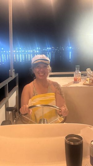 Smiling person in a captain’s hat steering a boat at night with marina lights reflecting on calm harbor water and a snack table nearby