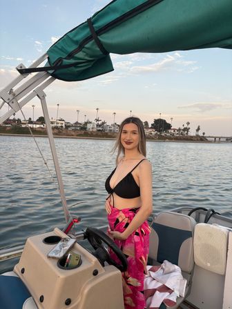 Smiling woman in a black bikini top with a pink floral sarong standing at the helm of a pontoon boat at sunset, calm harbor waters and palm-tree lined coastal homes in the background