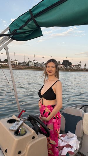 Smiling woman in a black bikini top with a pink floral sarong standing at the helm of a pontoon boat at sunset, calm harbor waters and palm-tree lined coastal homes in the background