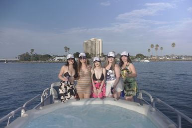 Five friends in swimsuits and matching trucker hats smiling on a boat with a bow hot tub, blue harbor water, palm trees and a coastal hotel skyline under a sunny sky.