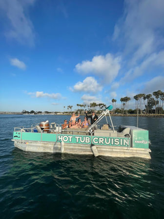 Group of friends in swimsuits relaxing on a turquoise hot-tub party boat cruising a sunny, palm‑lined coastal harbor under a bright blue sky.