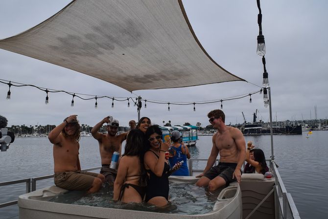 Friends in swimsuits laughing in a hot tub on a pontoon boat under a sunshade at a marina with string lights and moored boats