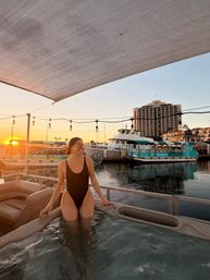Smiling person in a black one-piece swimsuit standing waist-deep in a hot tub at a marina sunset, string lights overhead and boats and waterfront buildings reflecting in the calm harbor water.