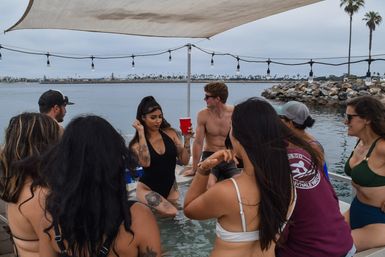 Group of friends at a boat party in a coastal bay under a shade sail and string lights, people in swimsuits socializing with a woman holding a red cup, rocky shoreline and palm trees in the background.