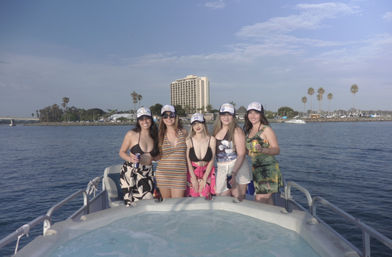 Five friends in swimsuits and summer dresses wearing matching caps and holding drinks, smiling on a pontoon boat with a hot tub in a sunny harbor lined with palm trees and a waterfront hotel.