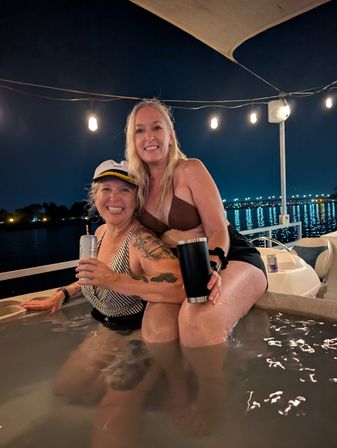 Two smiling women in swimsuits sharing drinks in a boat hot tub at night under string lights, with bridge and waterfront lights reflecting on the water