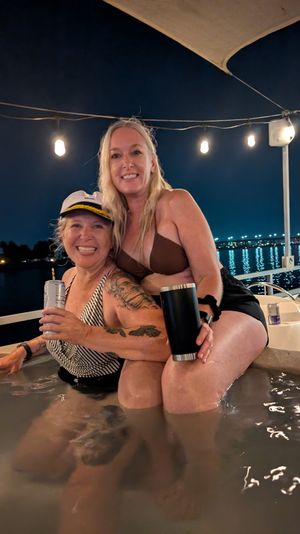 Two smiling women in swimsuits sharing drinks in a boat hot tub at night under string lights, with bridge and waterfront lights reflecting on the water