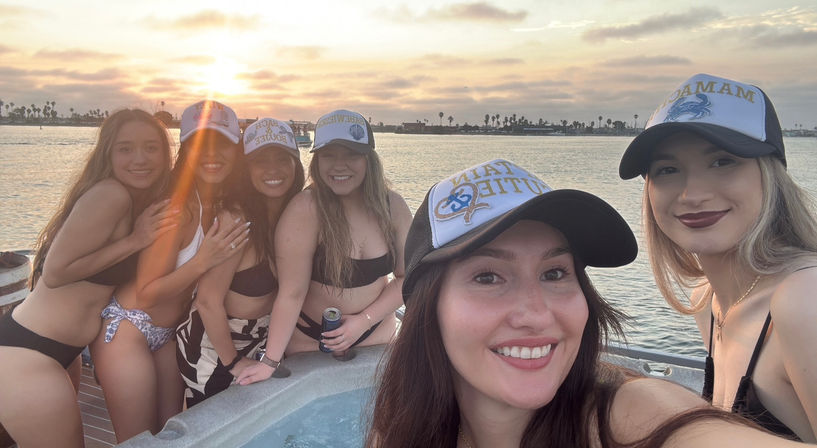 Six friends in bikinis and trucker hats posing around a boat hot tub during a golden sunset over a palm-lined coastal bay