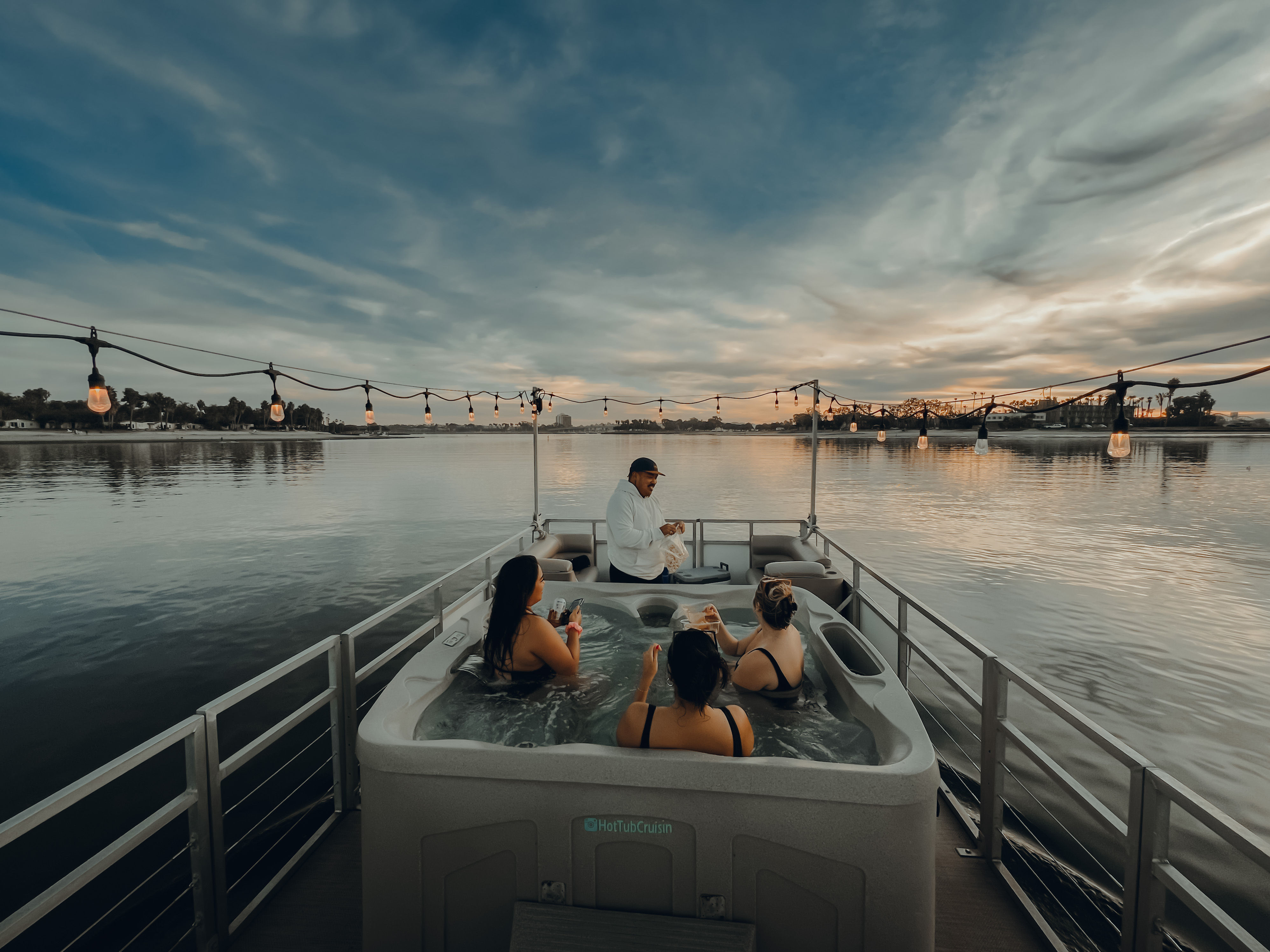 Three people soaking in a hot tub on a pontoon boat at sunset, a server offering drinks, string lights overhead and calm waterfront with dramatic sky