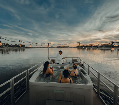 Three people soaking in a hot tub on a pontoon boat at sunset, a server offering drinks, string lights overhead and calm waterfront with dramatic sky