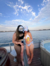 Two people in bikinis and trucker hats holding canned drinks on a boat deck with calm bay water, distant shoreline and blue sky with scattered clouds — summer coastal boating scene.