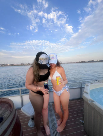 Two people in bikinis and trucker hats holding canned drinks on a boat deck with calm bay water, distant shoreline and blue sky with scattered clouds — summer coastal boating scene.