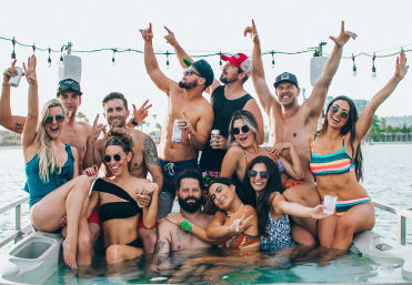 Cheerful summer boat party: a group of friends in swimsuits lounging in an onboard hot tub with drinks, string lights overhead and a waterfront shoreline in the background.