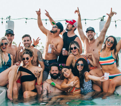 Cheerful summer boat party: a group of friends in swimsuits lounging in an onboard hot tub with drinks, string lights overhead and a waterfront shoreline in the background.