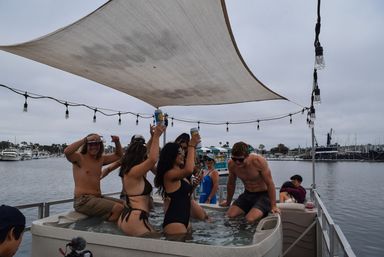Friends splashing in a hot tub on a pontoon at a marina, raising canned drinks under a fabric sunshade and string lights with boats and a cloudy sky beyond.
